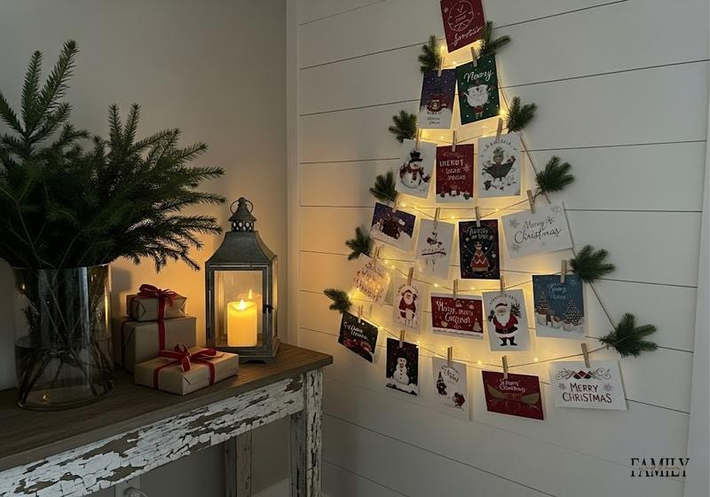 Christmas cards clipped on twine in a tree shape with fairy lights and evergreen accents.